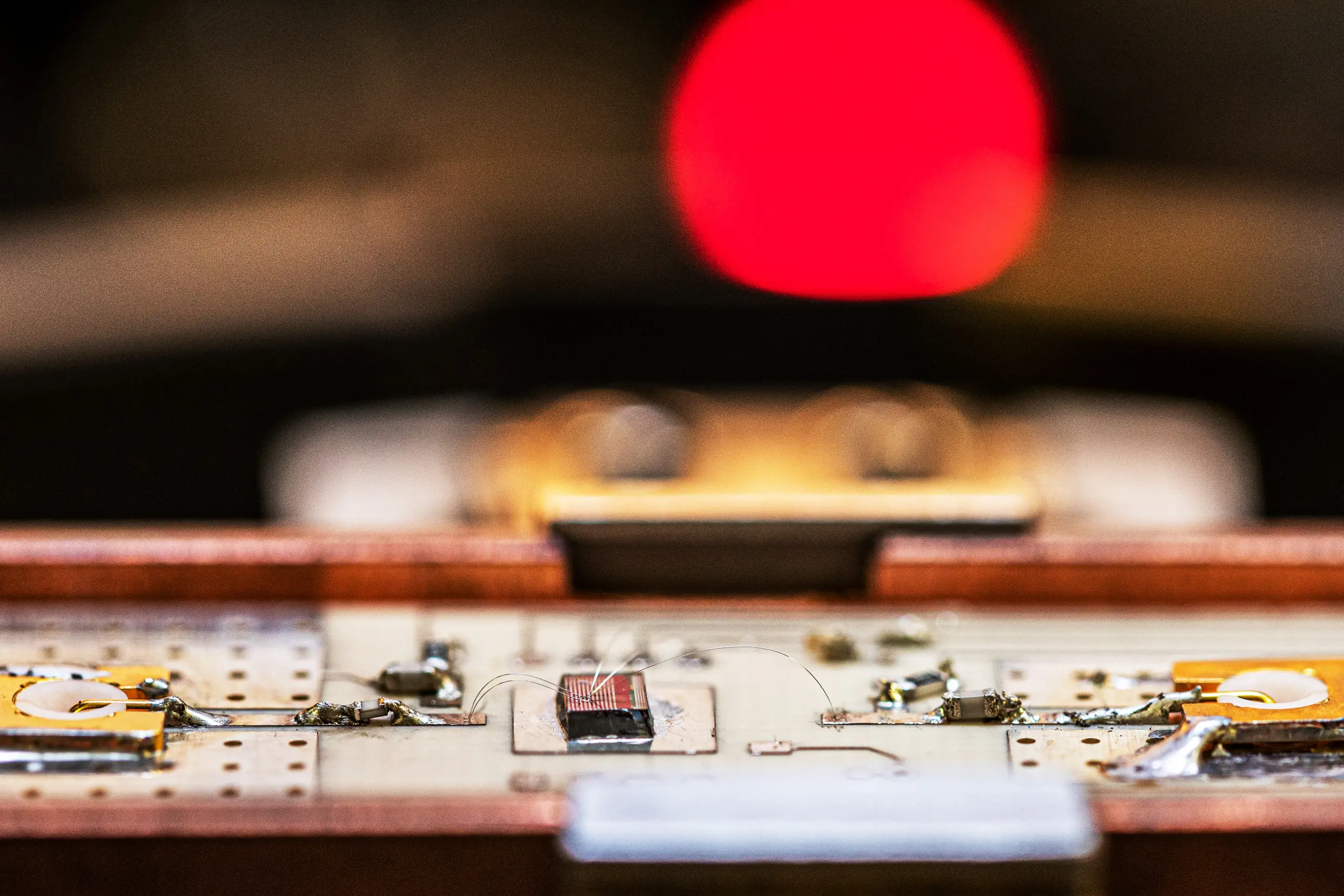 Close-up of an electronic circuit board with various components, focusing on the central part, against a blurred background featuring a prominent red element.