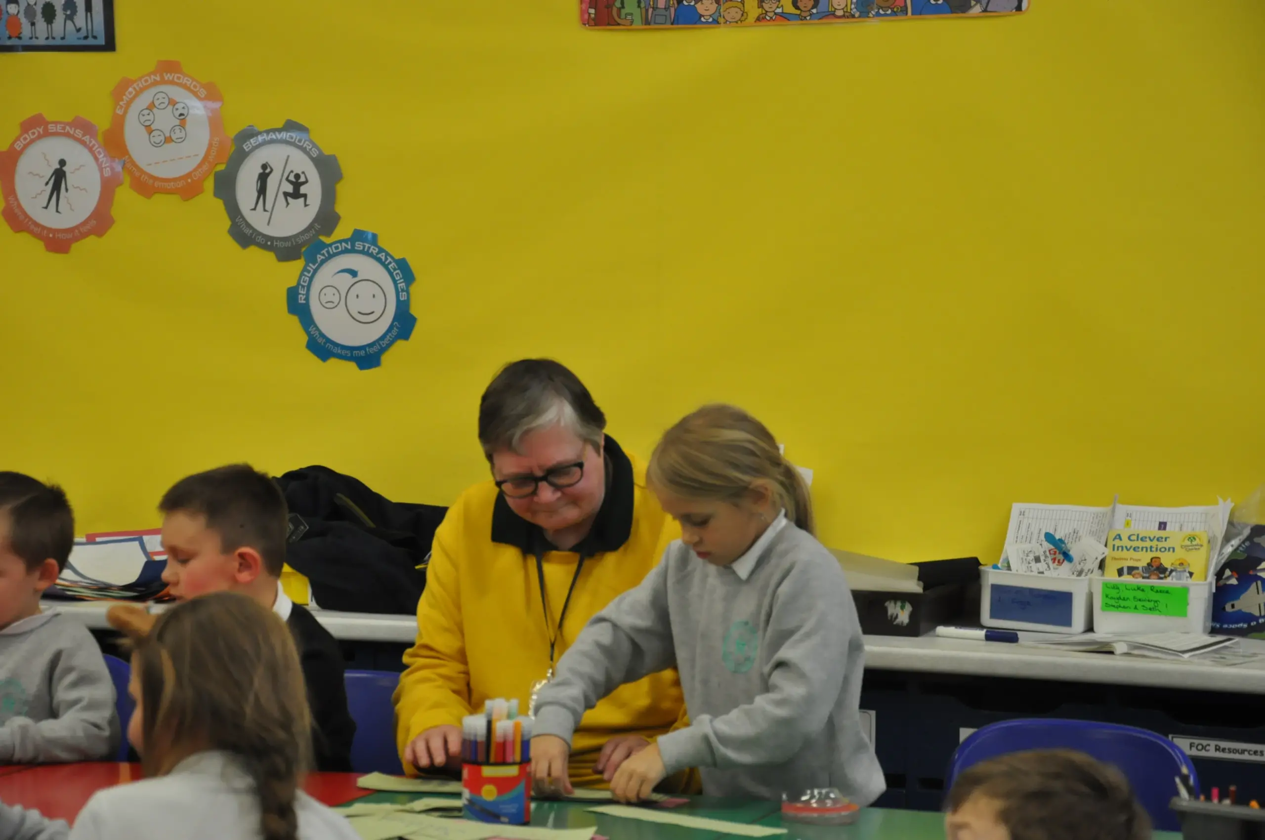 An adult in a yellow jacket assists a young girl with a craft project in a classroom, surrounded by other children.