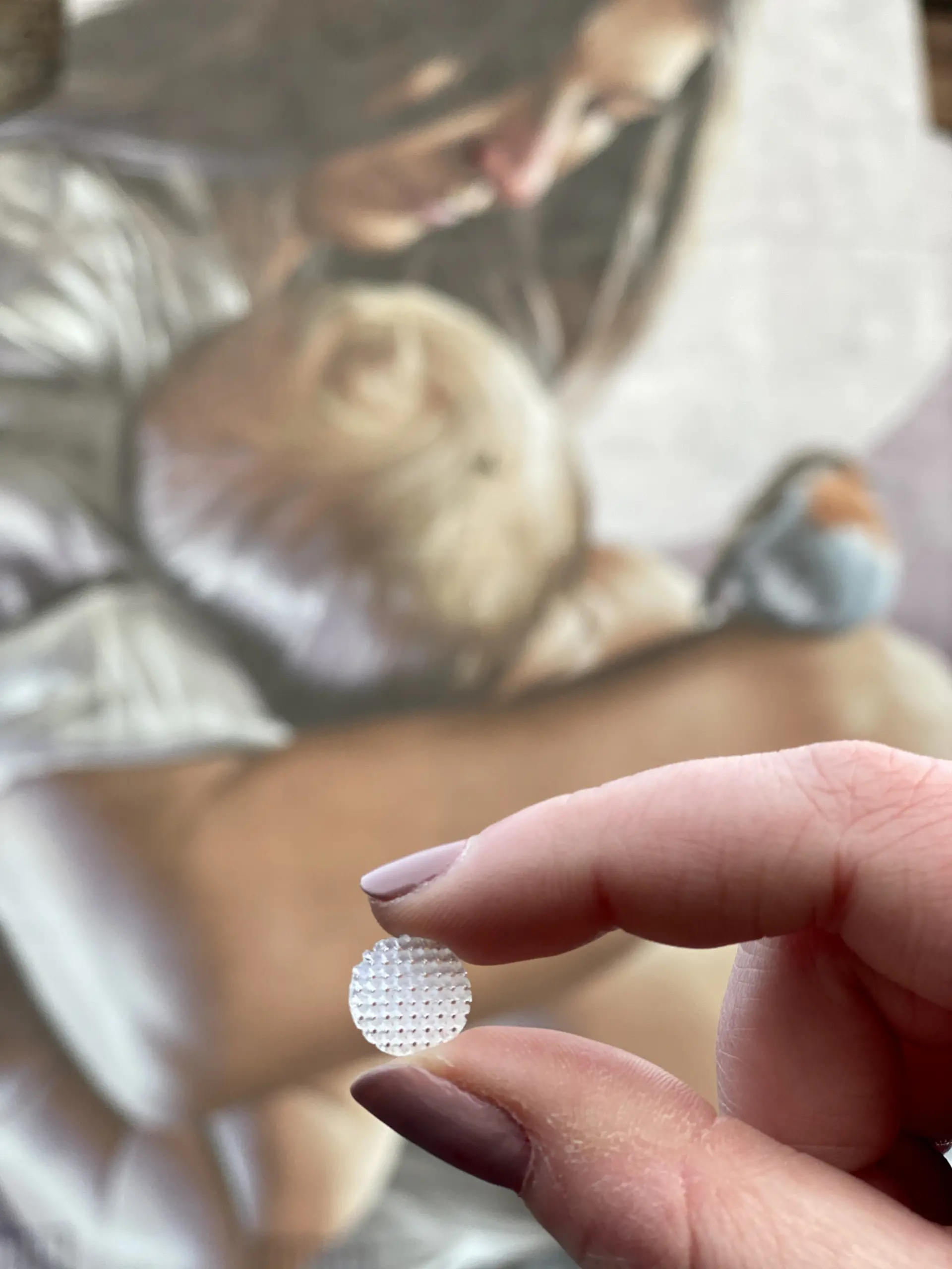 A hand holding a translucent circular object with a blurred mural of a woman holding a child in the background.