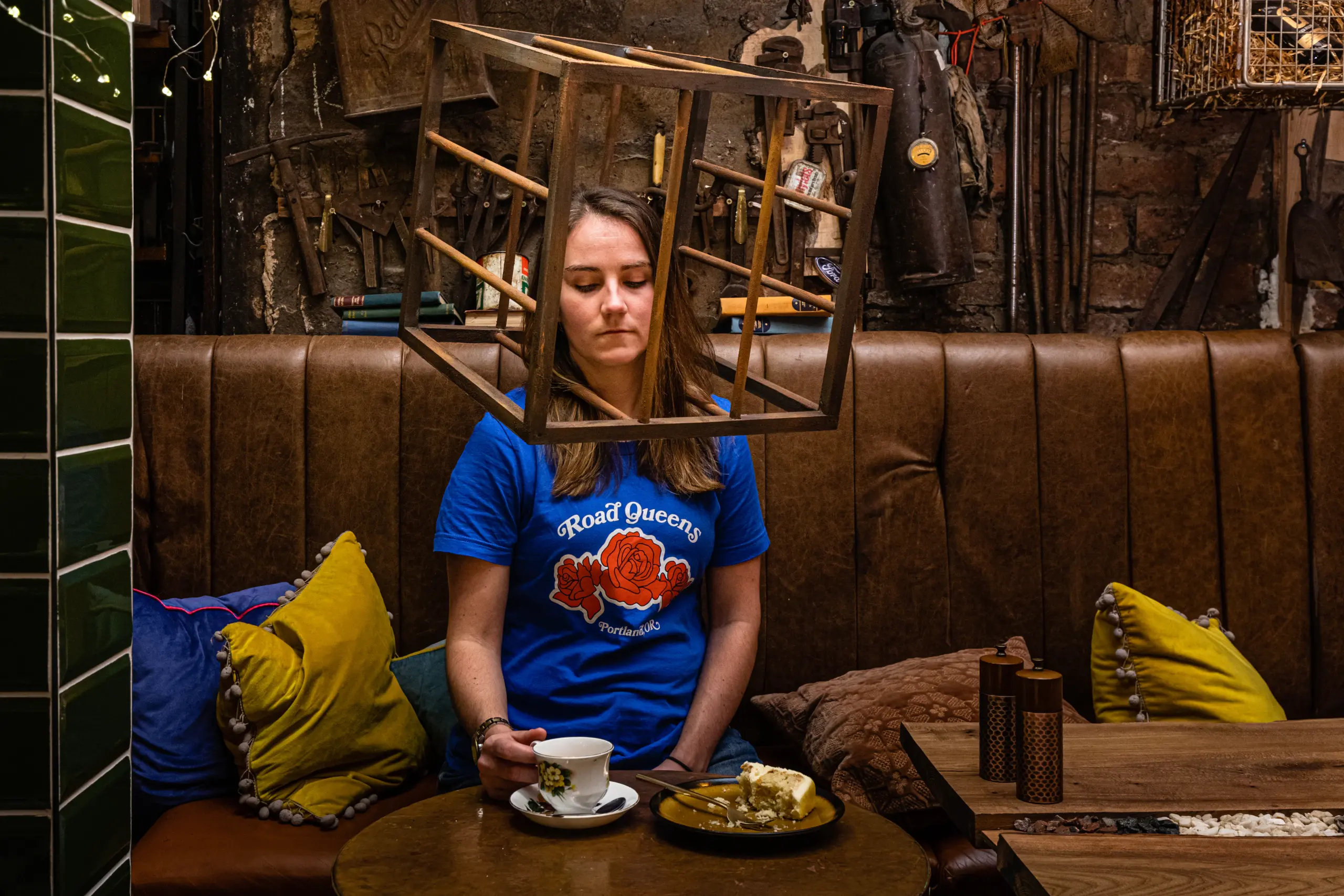 A woman in a blue shirt with "Road Queen" on it sits in a vintage cafe booth, holding a teacup, with a wooden cube frame suspended above her head.