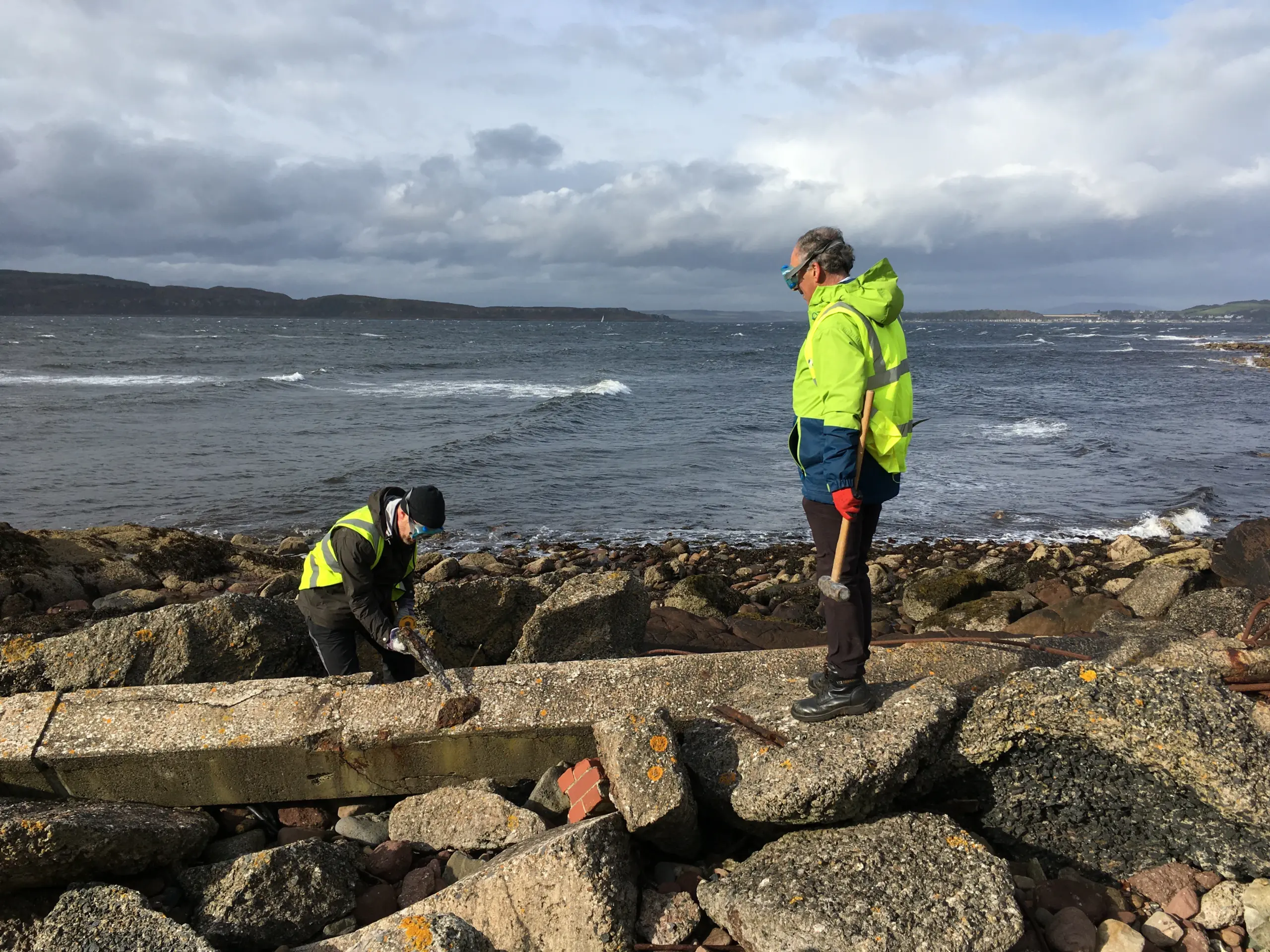 Two individuals in high-visibility jackets work on a rocky shoreline, examining a concrete structure. The sea and cloudy sky are in the background.