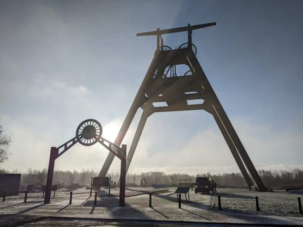 A large industrial structure with a wooden framework stands against a misty sky, with the sun partially visible behind it. A metal archway is in the foreground, casting a shadow on the ground.