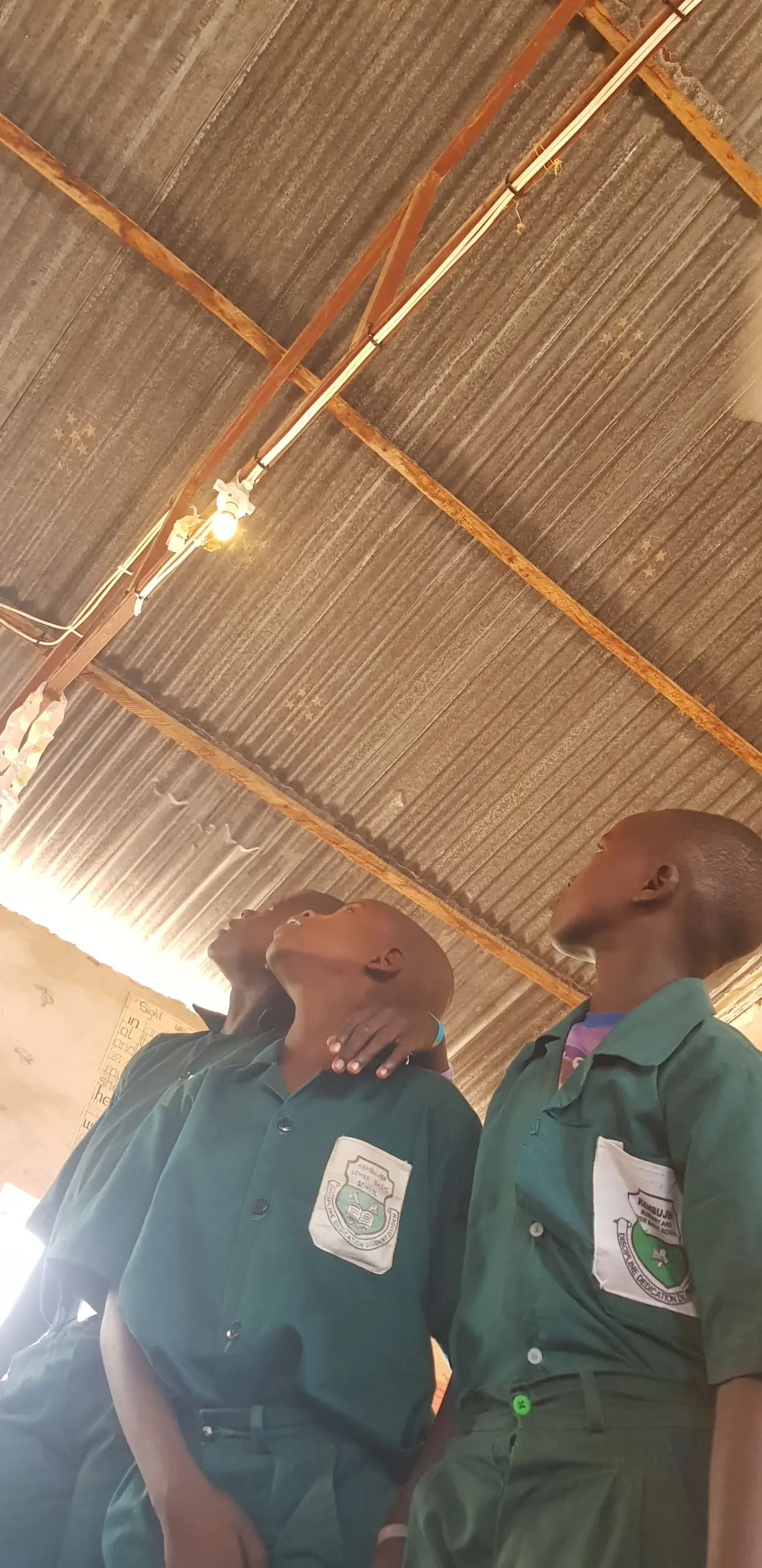Three students in green uniforms look up at the ceiling in a building with a corrugated metal roof. One student has his hand on another's shoulder.