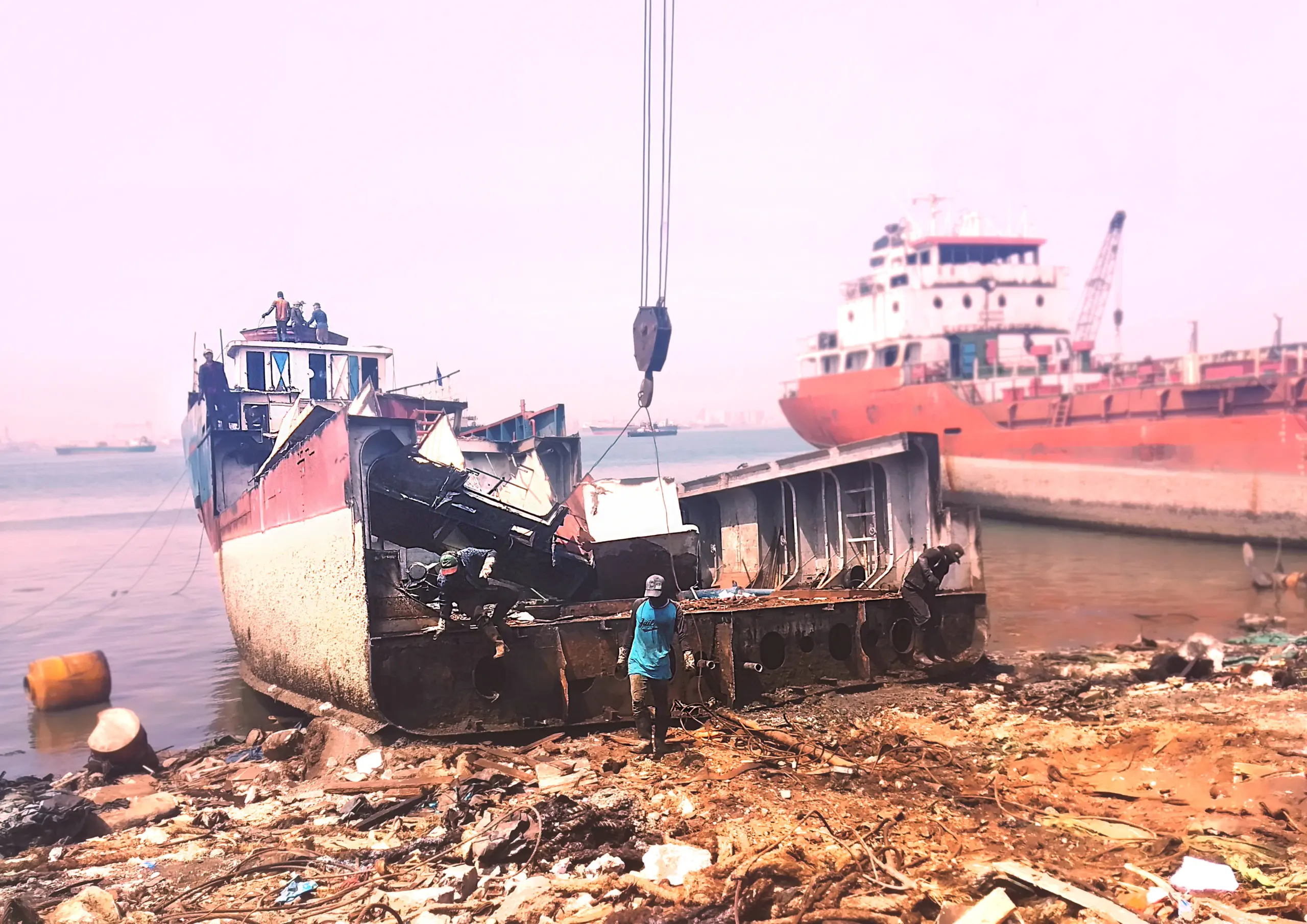 A partially dismantled ship sits on a polluted shore, with a crane lifting debris and a worker nearby. Another ship is docked in the background.